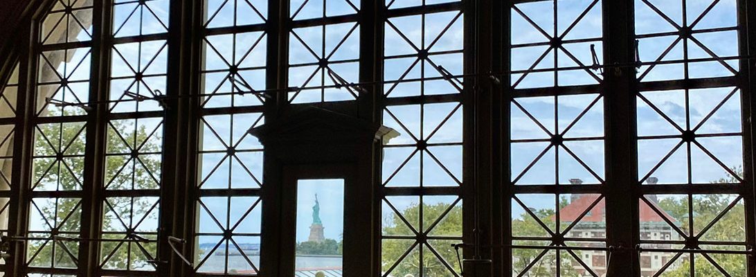 A photo of a large window at Ellis Island, the Statue of Liberty is seen in the distance